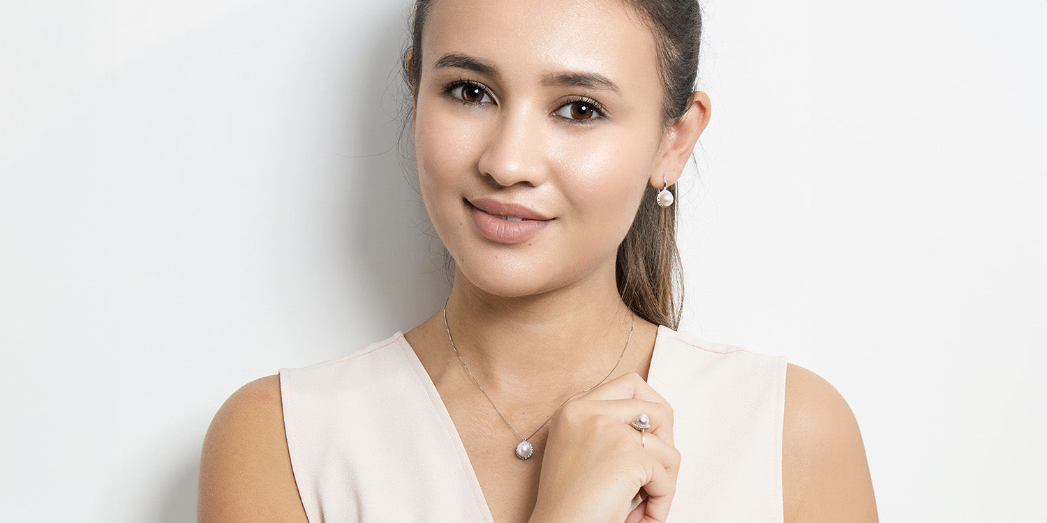 Lächelnde Frau trägt ein komplettes Schmuckset: goldene Halskette mit Perlen-Anhänger, passende Ohrstecker und Perlen-Ring, dazu ein beiges ärmelloses Top