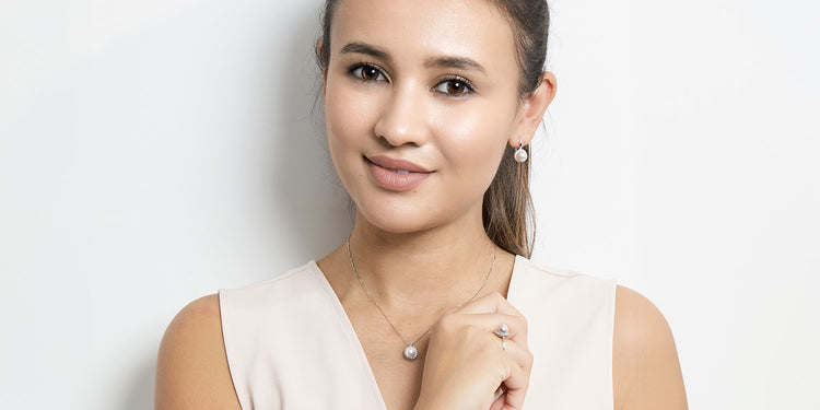 Lächelnde Frau trägt ein komplettes Schmuckset: goldene Halskette mit Perlen-Anhänger, passende Ohrstecker und Perlen-Ring, dazu ein beiges ärmelloses Top