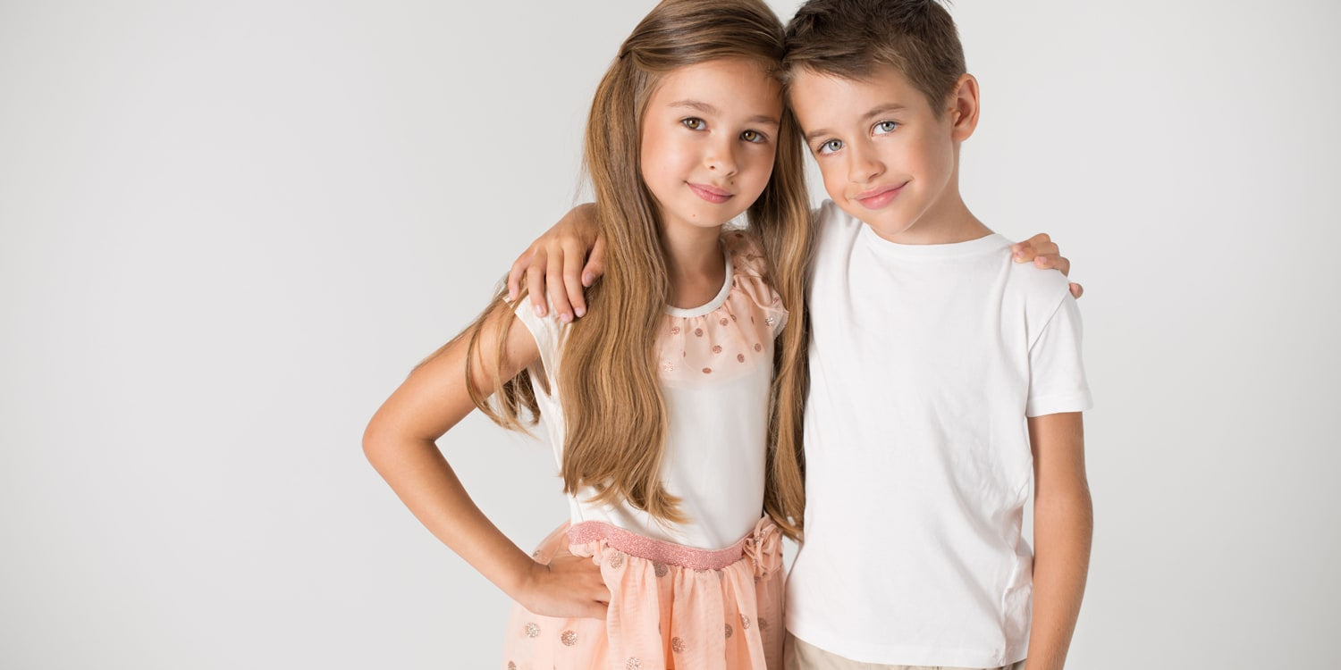 Zwei lächelnde Kinder umarmen sich - Mädchen mit langen blonden Haaren trägt rosa Kleid, Junge trägt weißes T-Shirt - Kinderschmuck