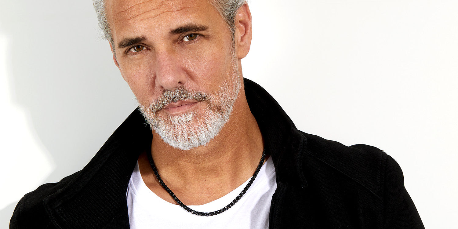 Eleganter Mann mit grauem Bart trägt schwarze geflochtene Halskette in schwarzer Jacke