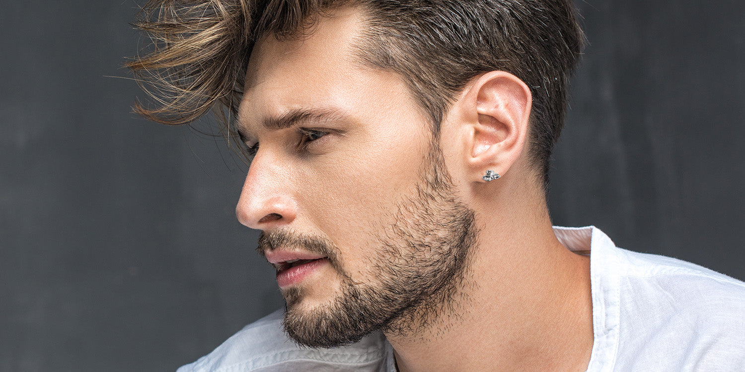 Junger Mann mit Bart in weißem T-Shirtträgt silbernen Ohrstecker mit Pistenbully-Motiv