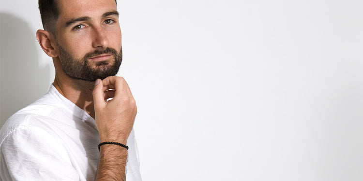 Mann mit Bart trägt schwarzes geflochtenes Lederarmband am Handgelenk zu weißem Hemd - maskuliner Herrenschmuck