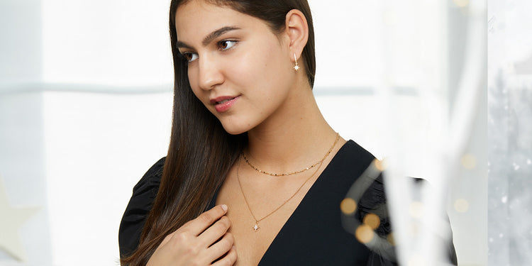 Elegante Frau mit langen braunen Haaren in schwarzem Top trägt goldene Layering-Halsketten mit Stern-Anhänger und goldene Stern-Ohrringe 