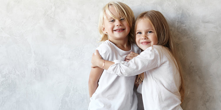 Zwei lächelnde Geschwister in weißen Shirts umarmen sich liebevoll - das Mädchen trägt ein Gravurarmband 