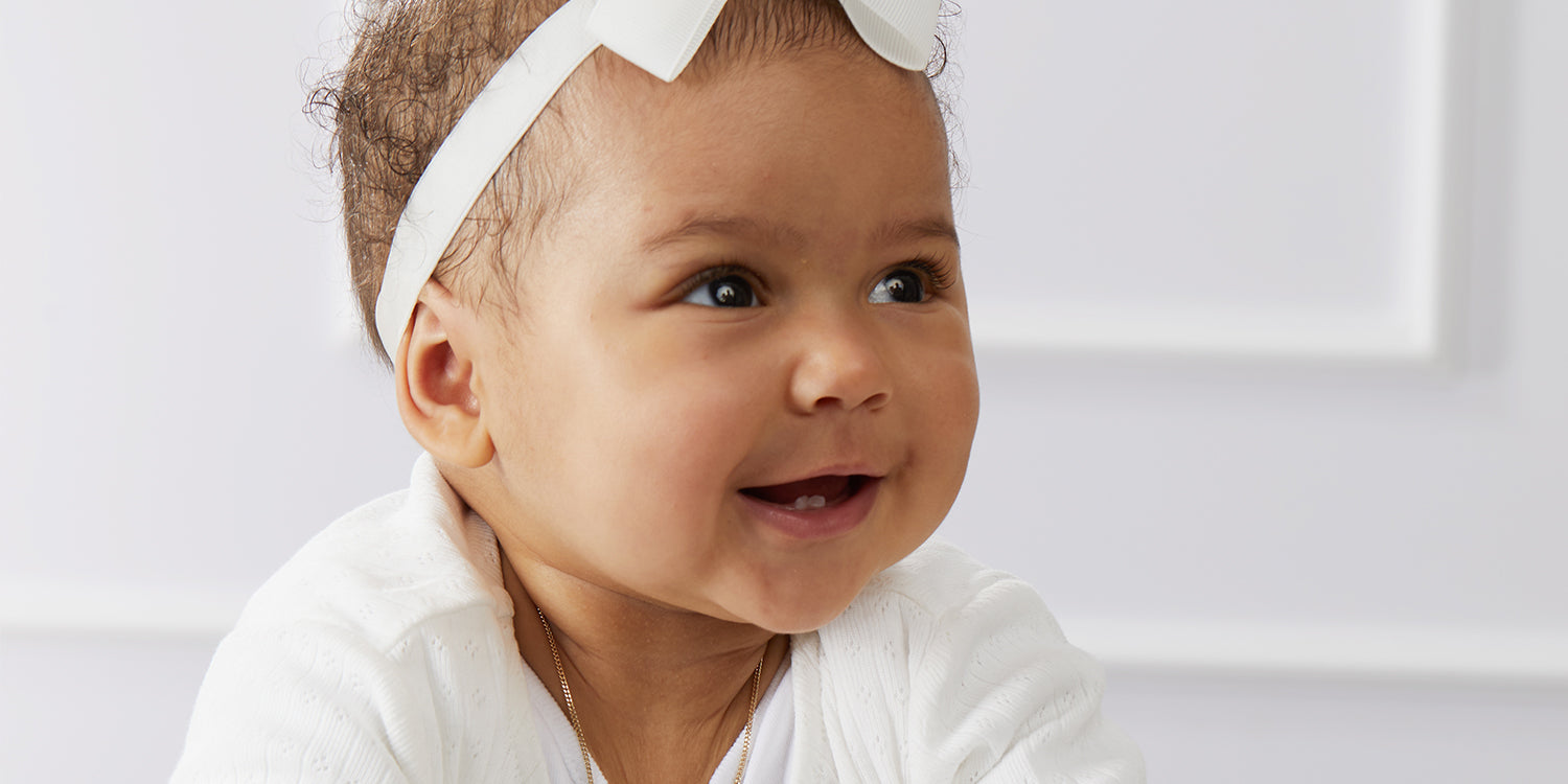 Lächelndes Baby mit weißem Haarband trägt eine zarte goldene Halskette zu weißer Kleidung - Schutzengel für Babys