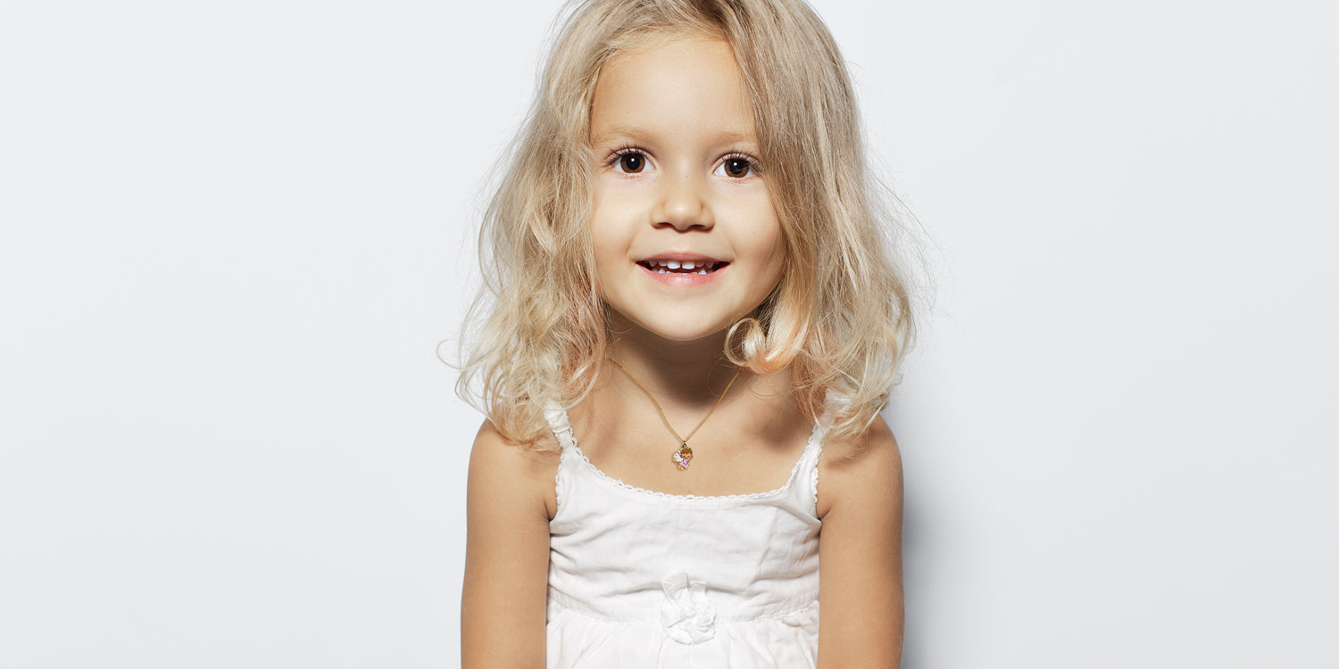 Lächelndes blondes Mädchen in weißem Kleid trägt zarte Halskette mit kleinem Schutzengel-Anhänger