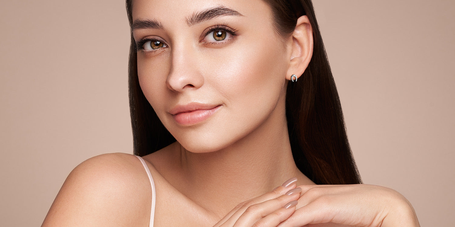 Junge Frau mit natürlichem Make-up trägt zarte silberne Ohrstecker in Form eines Hufeisens vor rosafarbenem Hintergrund