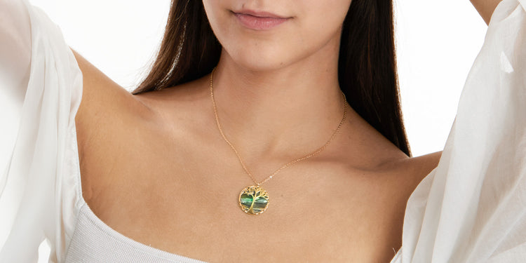 Frau mit langen braunen Haaren in weißem schulterfreien Top trägt goldene Halskette mit grünem Lebensbaum-Anhänger 