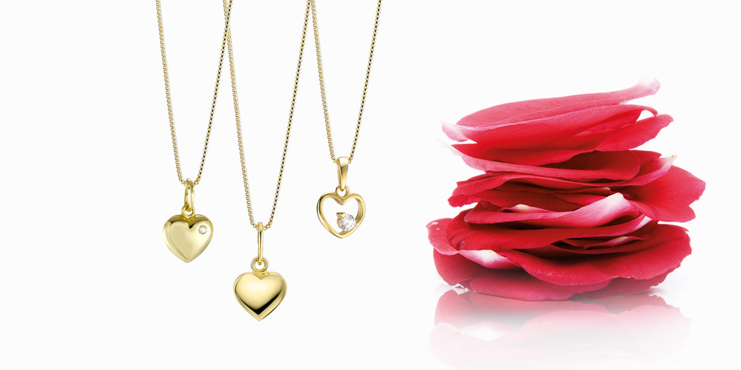 Drei goldene Halsketten mit Herz-Anhängern neben gestapelten roten Rosenblättern auf weißem Hintergrund