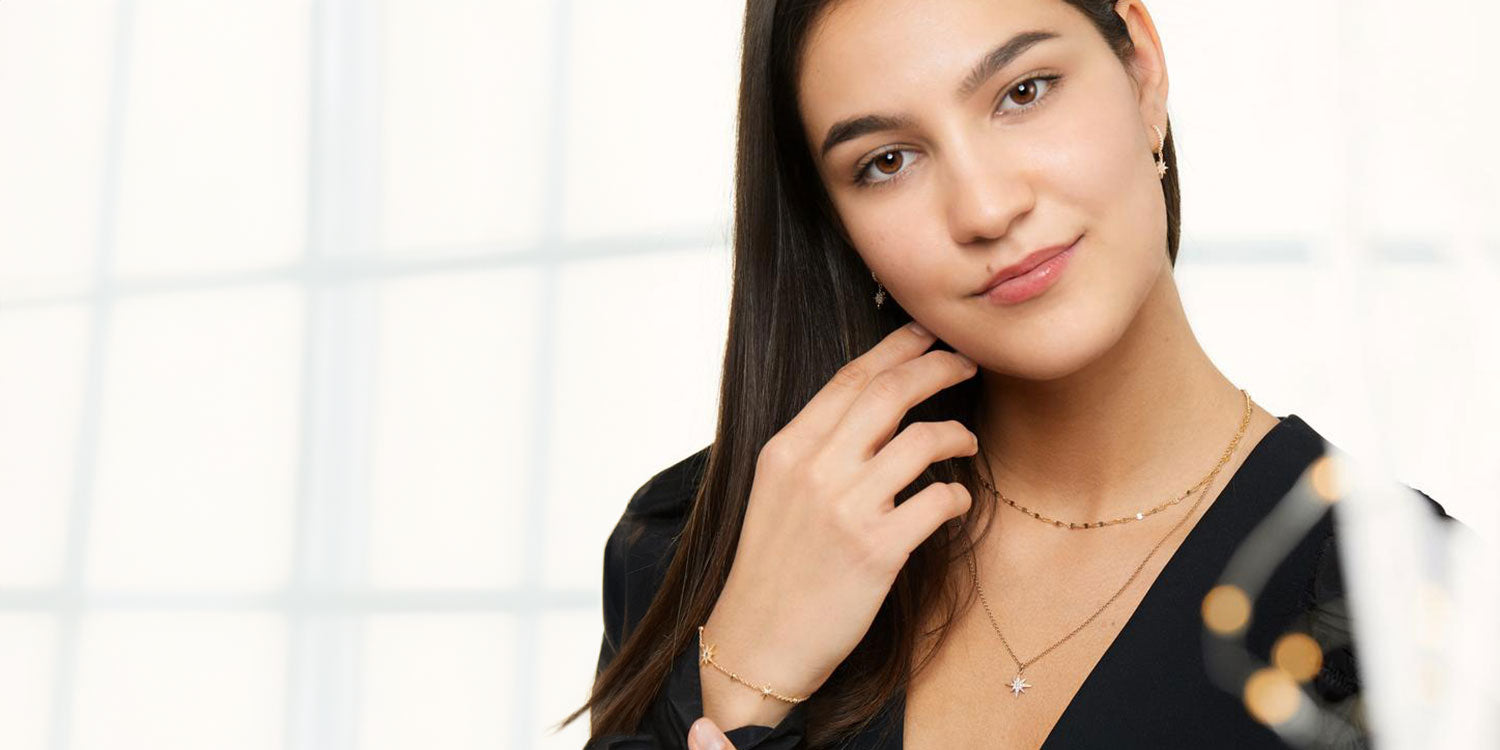 Lächelnde Frau mit langen braunen Haarenin schwarzem Top trägt goldene Layering-Halsketten mit Stern-Anhänger und goldenes Armband 