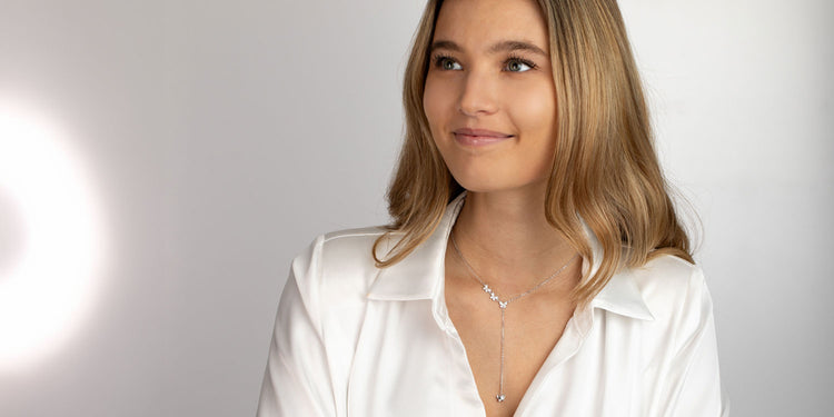 Frau mit blonden leicht gewellten Haaren trägt ein silbernes Y-Collier zu einer weißen Bluse, lächelnd vor hellem Hintergrund