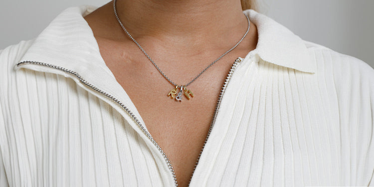 Frau trägt silberne Halskette mit goldenen Buchstaben-Anhängern über weißer gerippter Bluse mit V-Ausschnitt
