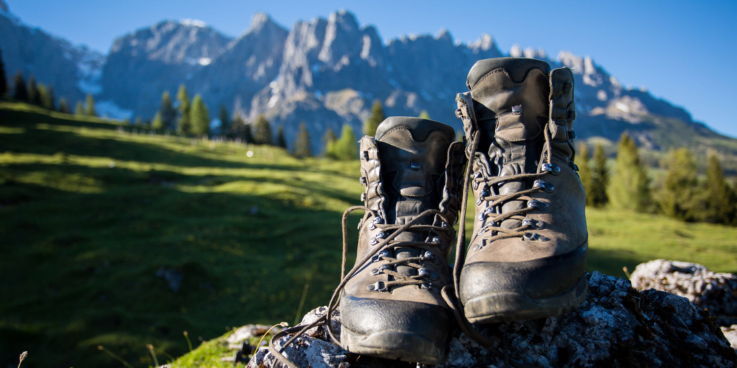 Braune Wanderstiefel auf einem Felsen vor alpiner Berglandschaft mit grünen Wiesen und schneebedeckten Gipfeln