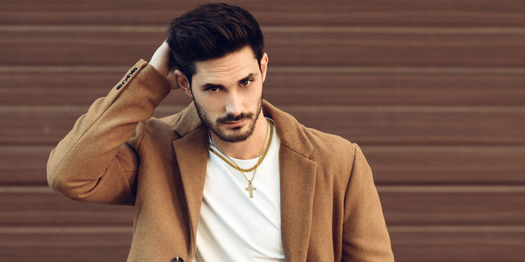 Stilvoller Mann mit mehrlagigem Goldschmuck, trägt eine kräftige Goldkette und Kreuzanhänger, gekleidet in einem kamelfarbenen Mantel über weißem T-Shirt