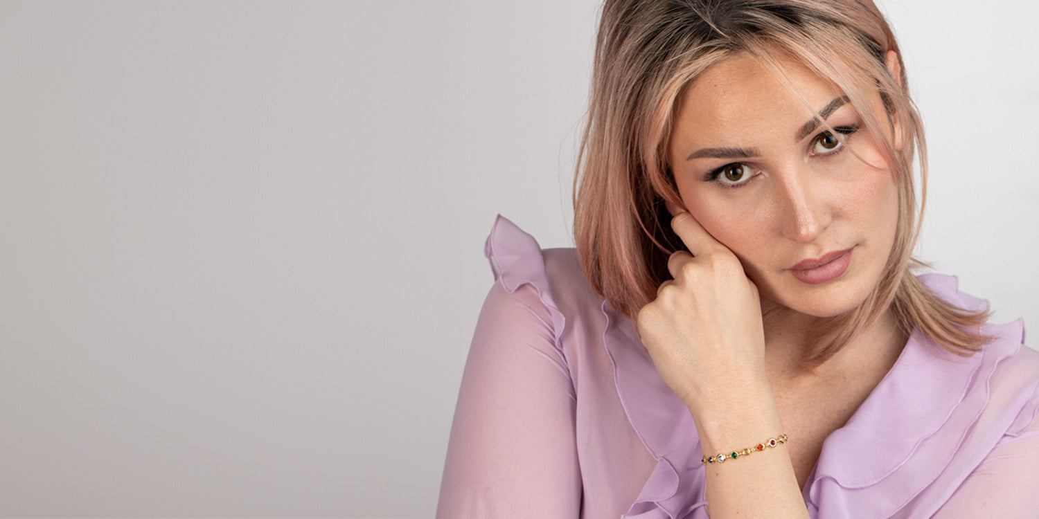 Blonde Frau in lila Rüschenbluse trägt zartes goldenes Armband mit bunten Edelsteinen