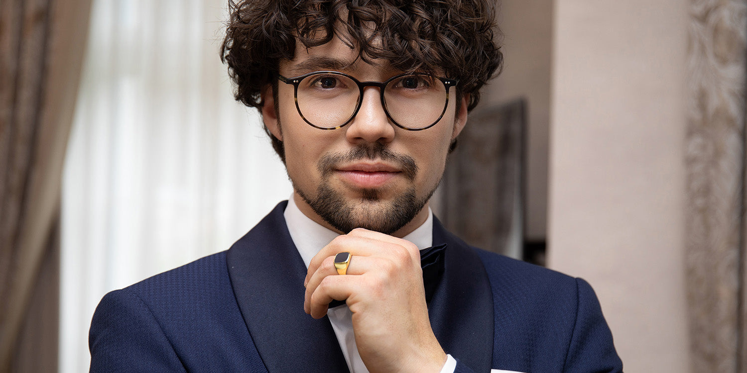 Eleganter Mann mit lockigen Haaren und Brille in dunkelblauem Anzug trägt goldenen Siegelring 